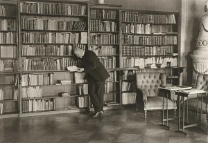 Tomáš Garrigue Masaryk se sklání nad otevřenou knihou u vysokých knihoven, vpravo čalouněná křesla, stolky a kachlová kamna