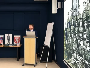 Historik Tomáš Jakl u řečnického pultu, vedle velkoplošná dobová fotografie vojáků