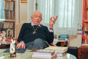 Petr Piťha v kněžském oděvu s křížem sedí v pracovně s knihovnou