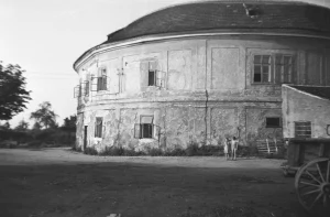 Černobílá fotografie oprýskané válcové budovy s okny a vozem v popředí
